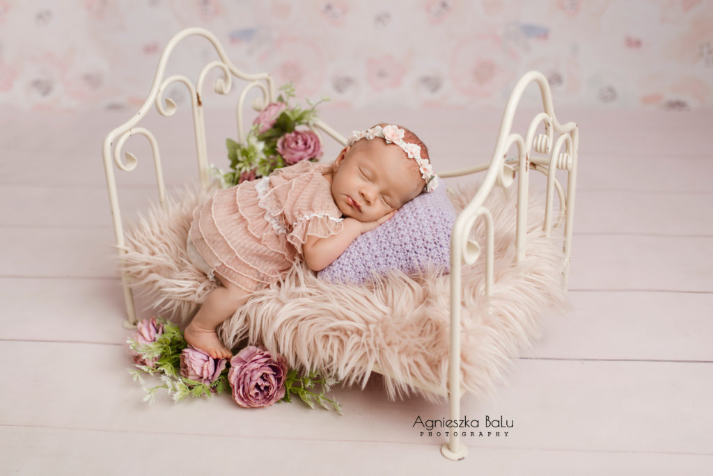 Das Babymädchen liegt auf dem Mädchenbett. Ein Lilakissen, rosa Decke und die Blumen bilden eine schöne Mädchenkulisse.