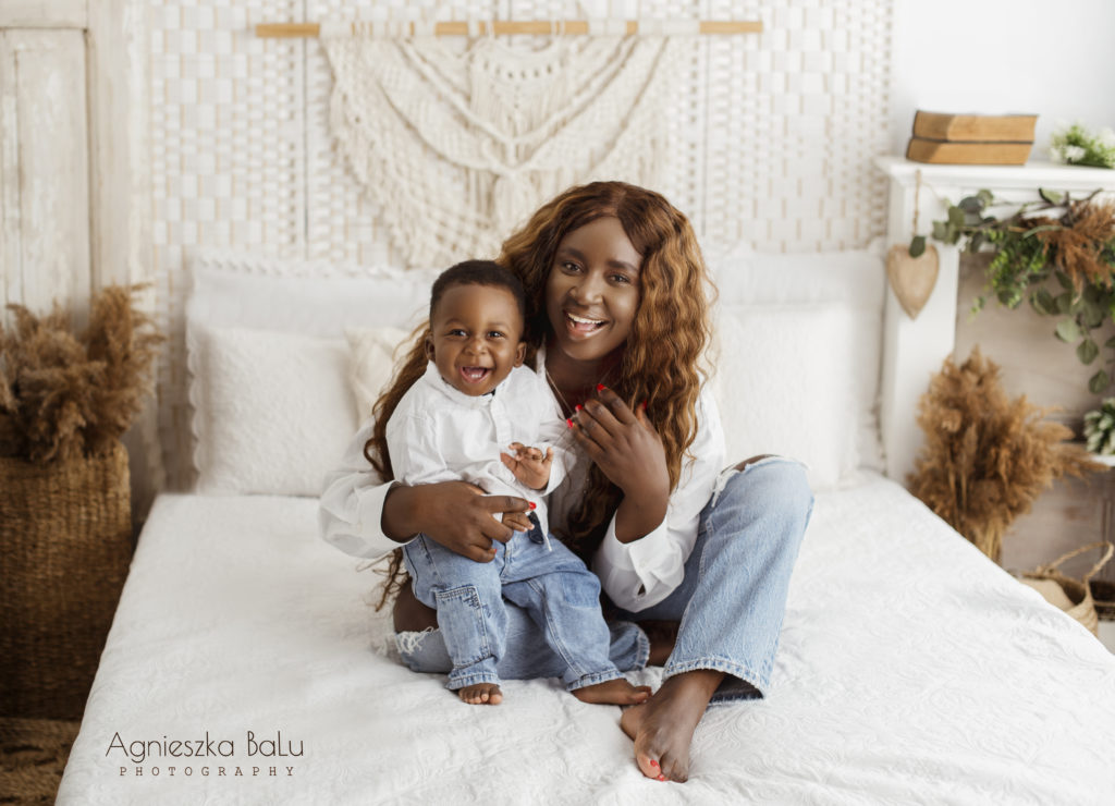 Die dunkelhäutige Frau halt das dunkelhäutige Babyjunge auf dem Schoß. Die Nigerianer sitzt auf dem Bett und hat die Jeans und weisses Hemd an. Die lächelt und zeigt Zähne. Das Papasgrass steht im Hintergrund und macht den Boholook.