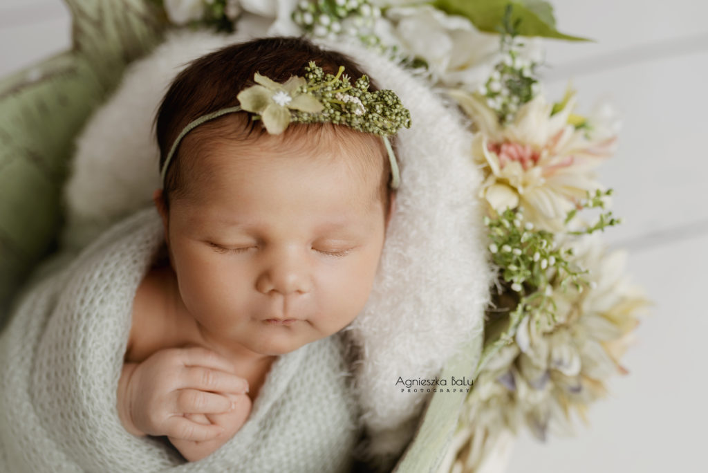 Das Baby liegt zwischen hellen Blumen. Das Mädchen trägt passender Stirnband. Die Hände sind verschränkt.
