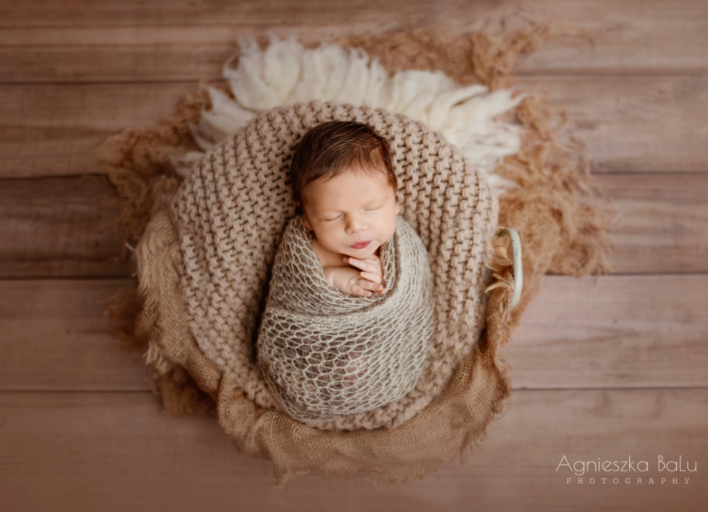 Das gepuckte Neugeborene liegt auf vielen bräunlichen Decken auf dem Holzbogen. Die Farben passen sehr gut zueinander und erstellen eine natürliche, zeitlose Fotografie.