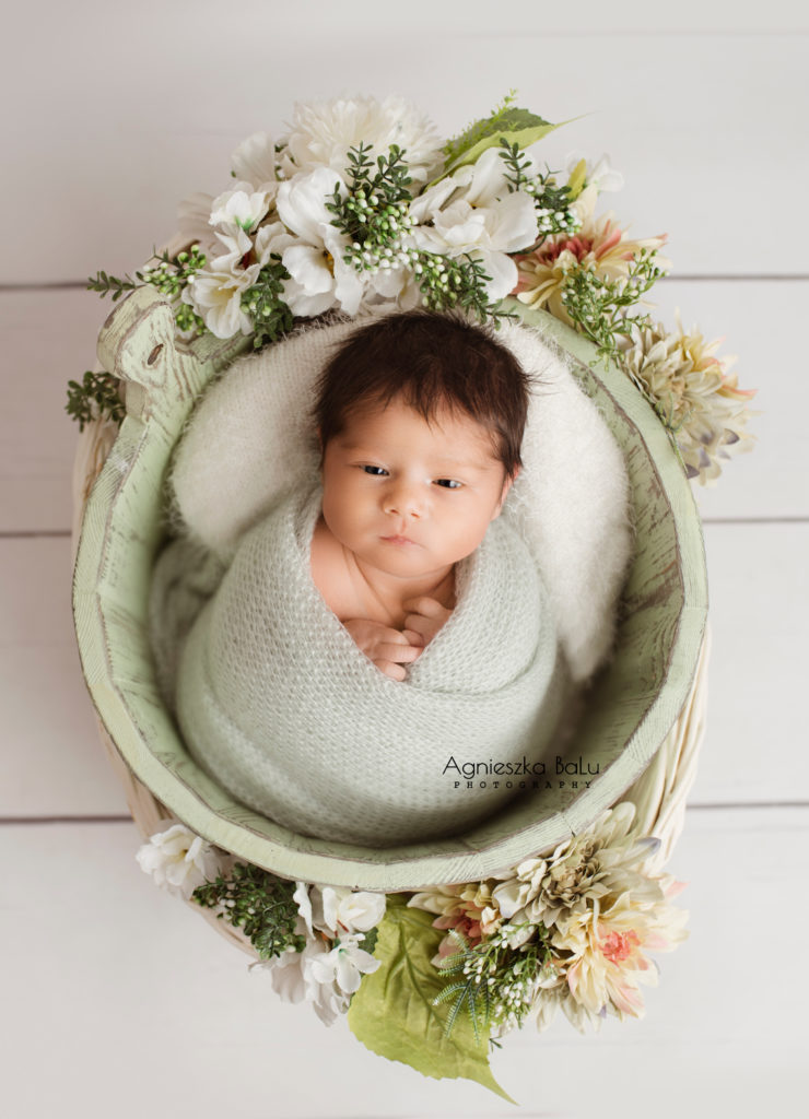 Das von grünen Tuch gepucktes Baby liegt in einem grünen Eimer. Daneben befinden sich viele grünen Blumen und der weiße Boden.