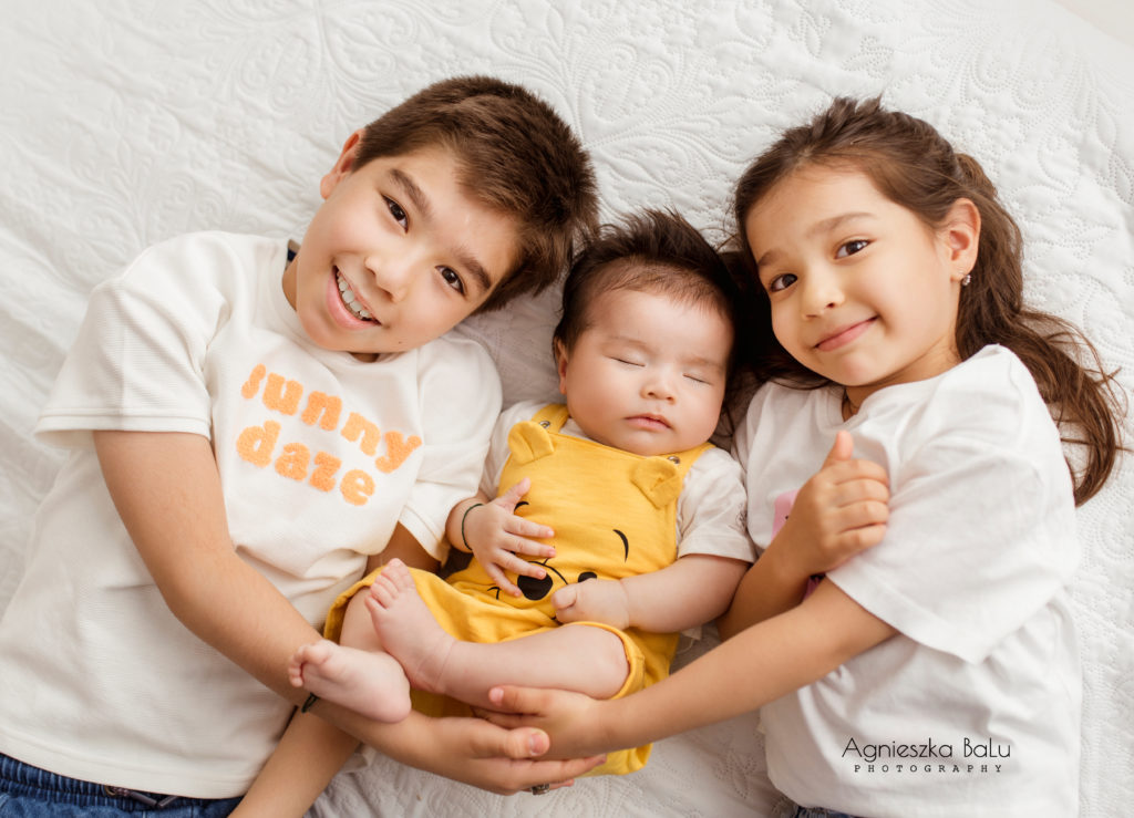 Die Geschwisterfotografie von einem Bruder und zwei Schwester. Die Kinder liegen eng zueinander und halten das Baby, das in der Mitte liegt.