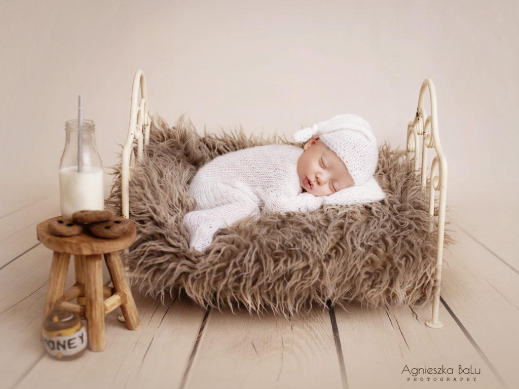 Das Baby liegt auf die braune Decke ist trägt weise Hose und weise Mütze. Daneben liegt ein kleines Tischen mit der Milch und Kuchen.