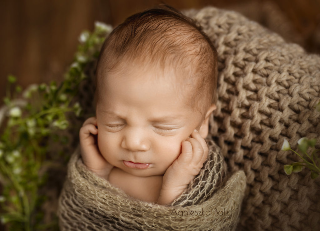 Die Nahaufnahme von dem Neugeborenen, die in der bräunlichen Farben abfotografiert wird. Das Baby hält die Händen neben dem Gesicht.
