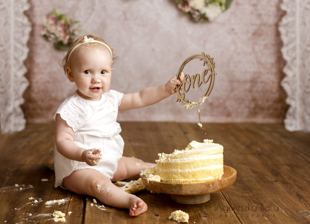 Das lachende Mädchen sitzt neben der Torte. Der Holzboden und die weißen Gardinen befinden sich im Hintergrund.