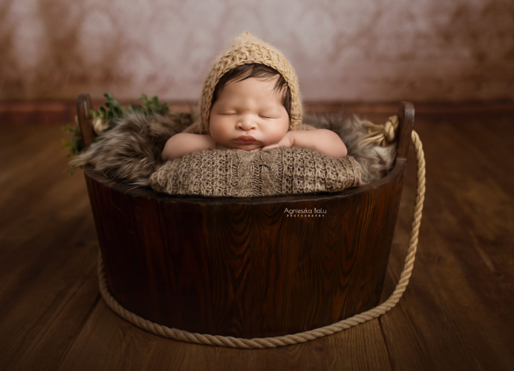 Die Babyfotografie von der Junge, die eine braune Mütze trägt und liegt in einem Holzeimer auf dem Holzboden. Die Requisitzen machenen einen Vintagelook.