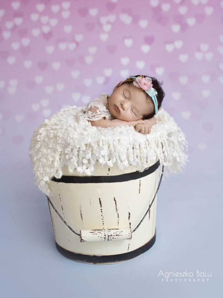 Ein Neugeborene liegt in einem beigen Eimer auf die weißen Decke und schläft. Der rosa-blaue Hintergrund passen zu dem Rosastirnband. Das Bild macht ein Mädchenlook.