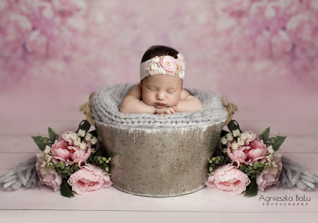 Das Neugeborenen liegt auf der grauen Decke im Eimer. Daneben lieben die Rosen. Im Hintergrund befinden sich auf die geschwollenen Rosenkonturen. Das Baby hält die Händchen unter dem Kopf. Das Bild ist eine wunderschöne Komposition.