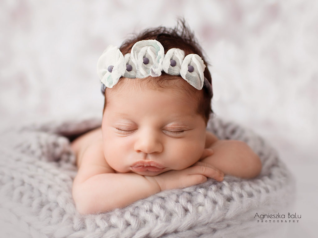 Das Neugeborene schälft auf dem Bett. Die graue Decke, ein graues Stirnband mit den Blumen und grauer Hintergrund passen zueinander.