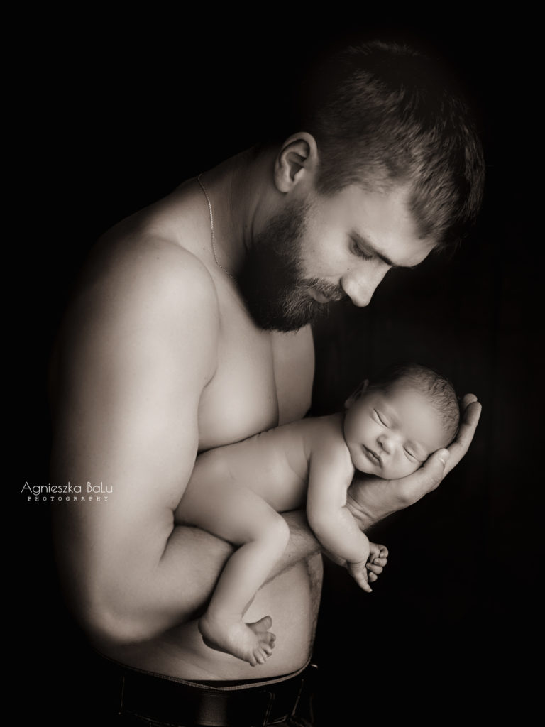 Der halbnackte Papa hält dein kleinen Sohn auf dem Arm. Das schwarz-weißes Bild zeigt wie klein ist das Baby. Die universelle Pose ist zeitlos und gehört zum Kanon der Neugeborenenfotos.