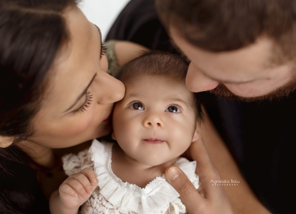 Die frisch gebackenden Elten küssen das Neugeborenen. Das Baby guckt in den Elternsaugen. Das Portraitbild ist herzberührend.