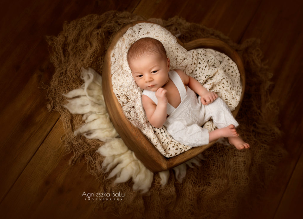 Das Neugeborenen liegt in einem Korb in Form eines Herzens. Das Herz ist braun und ist aus Holz. Das Baby hat helle Hosen und liegt auf der beigen Decke. Die Vintige Hintergrund ist braun. Das Baby liegt seitlich. Die natürliche Pose lässt das Baby sehr süß aussehen.
